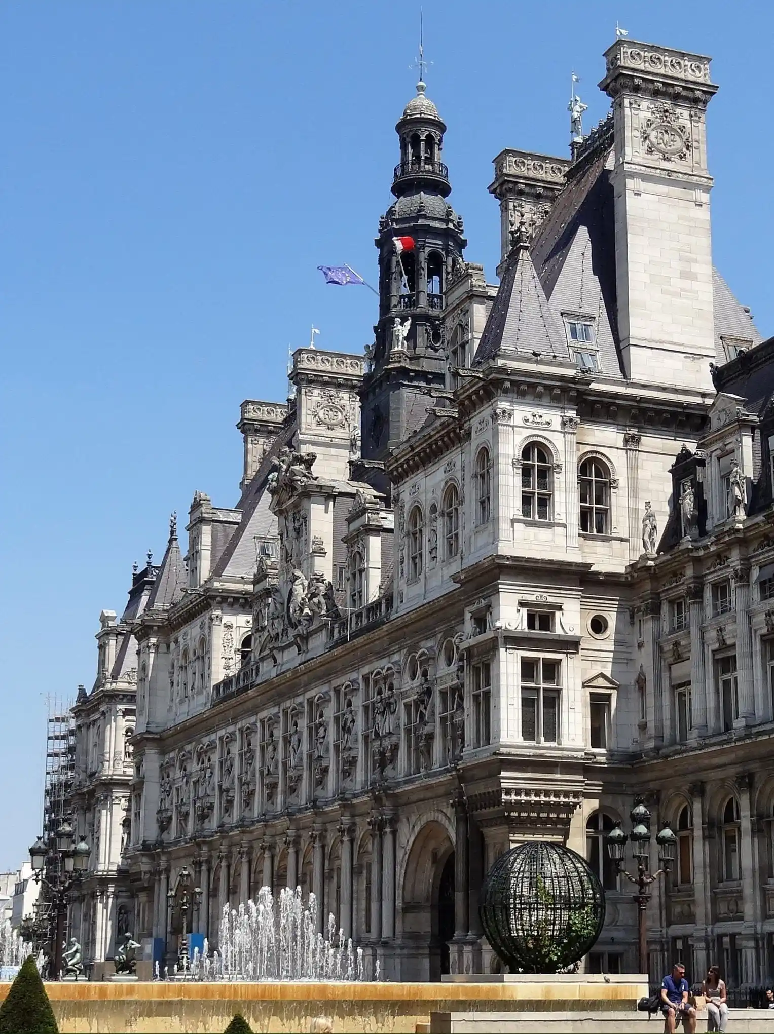 Façade sculptée d’un hôtel de ville avec clochetons, drapeaux et fontaine devant.